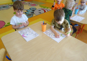 Dwójka dzieci koloruje zaszyfrowane według kodu ilustracje dinozaurów.